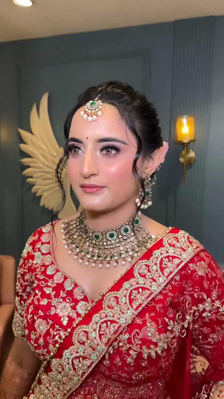 A bride in a heavily embroidered red lehenga with a kundan and emerald choker and a maang tikka, standing beside a wall sconce and a gold wing sculpture.