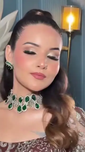 A close-up of a woman with soft smoky eye makeup and an emerald and diamond choker, eyes lowered, a lit wall sconce behind her.
