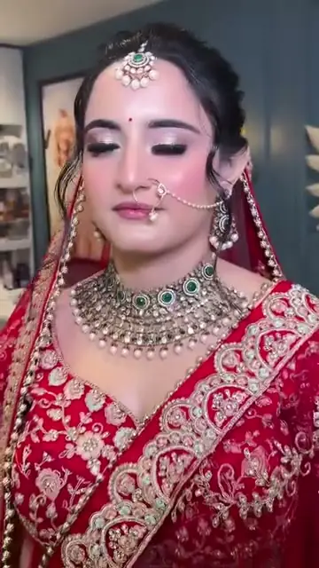 A bride in red wearing a nose ring joined by a chain to her hair, a kundan choker and a maang tikka.