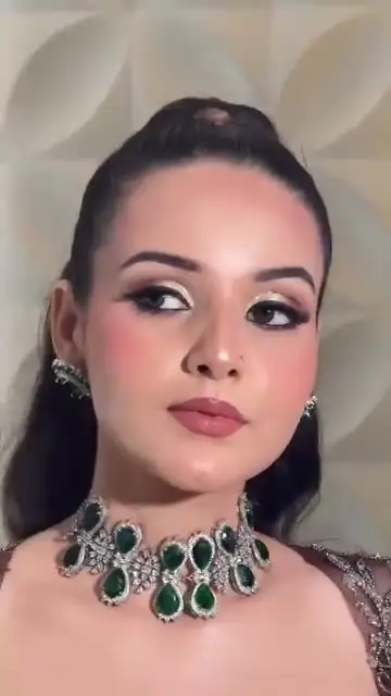 A woman with glam makeup and long loose waves wearing an emerald and diamond choker, a pale sculpted wall behind her.